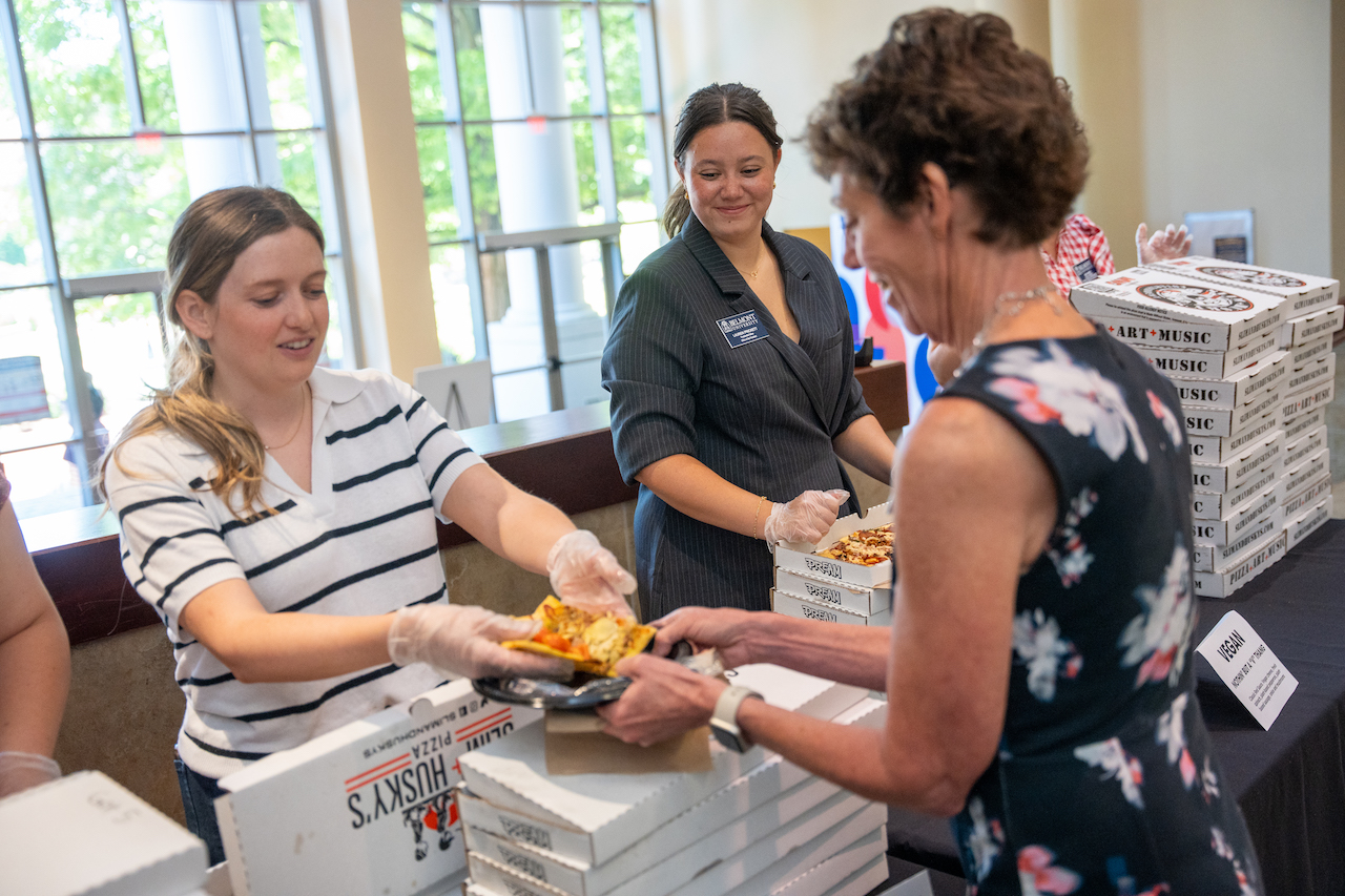 Female volunteers in black and white stripes smiles and hands out pizza to another woman in a black floral dress