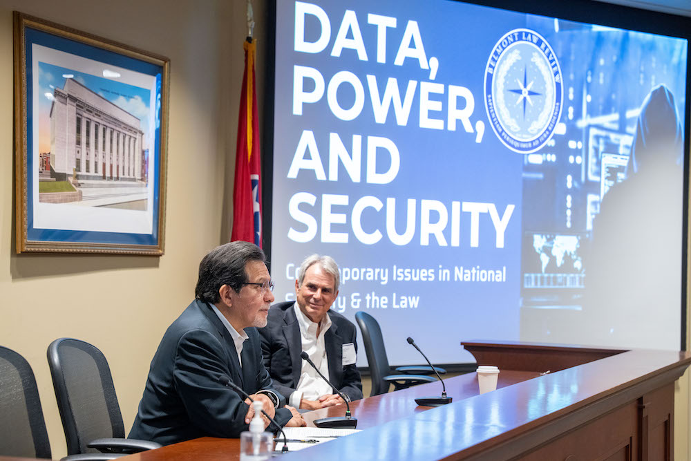 John Bellinger and Alberto Gonzales sit at a table in front of a large projected screen displaying symposium theme
