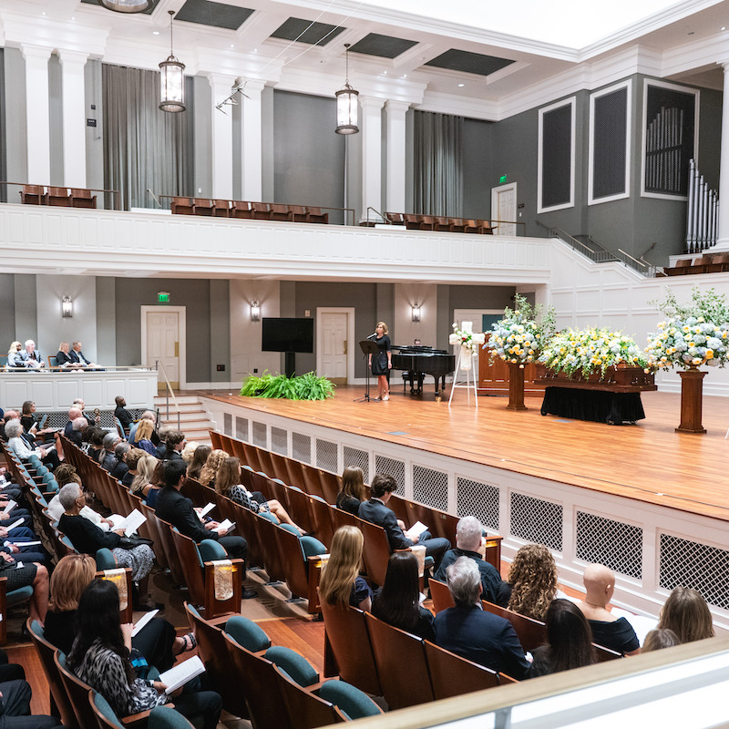 Audience seated in McAfee Concert Hall during Barbara Massey Rogers’ celebration of life, with a speaker at the podium and floral arrangements beside the casket on stage.