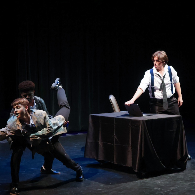 one actor holding another as he reaches across the stage with a third standing in the background behind a desk