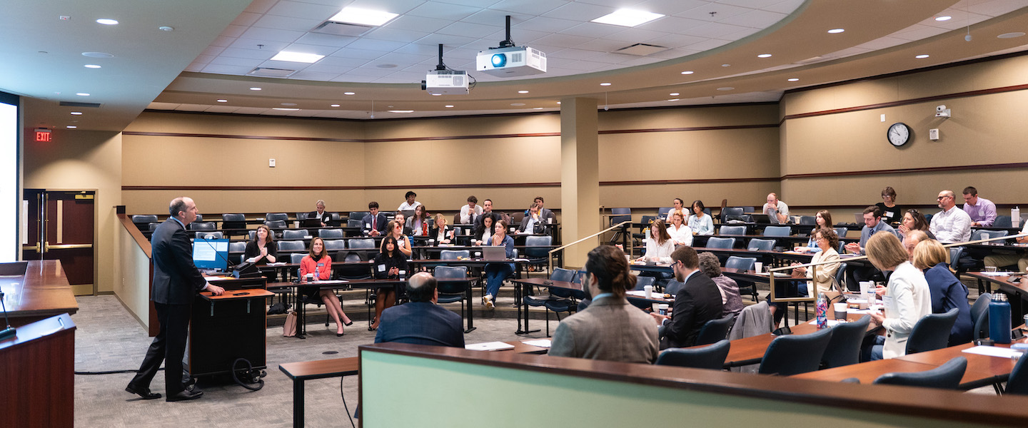 speaker presents to an audience seated in a tiered lecture hall.