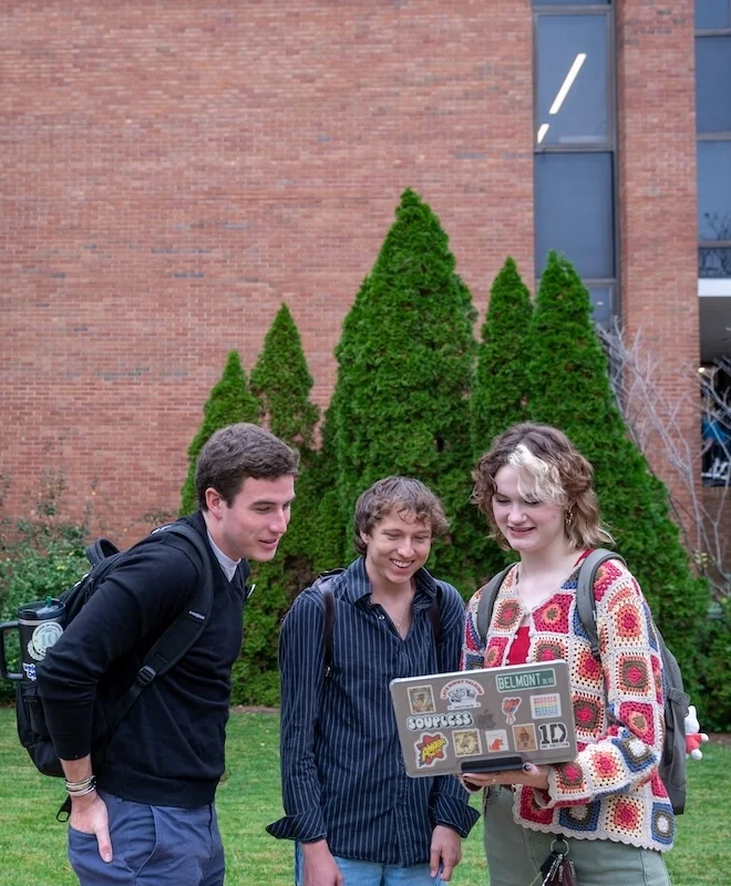 Three Belmont University students stand outside Hitch Building, smiling as they look at a laptop covered in stickers, including one that reads “BELMONT.”