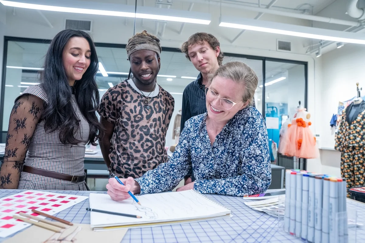 Fashion merchandising and design students at Belmont University gather around an instructor who sketches in a notebook, surrounded by fabric swatches and markers in a studio setting.