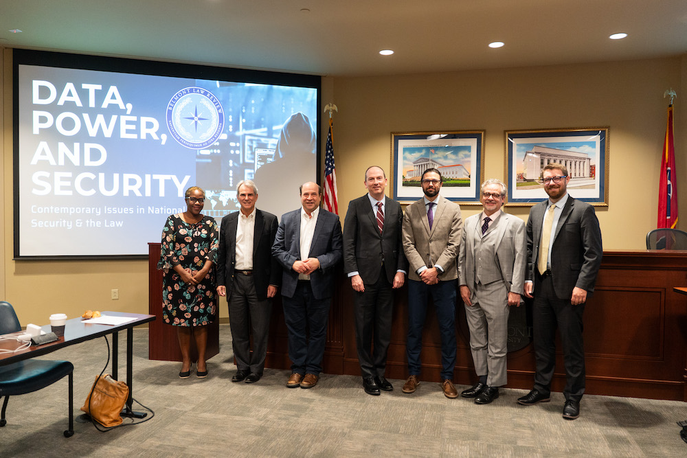 Symposium speakers stand side by side in front of a screen that reads the symposium theme