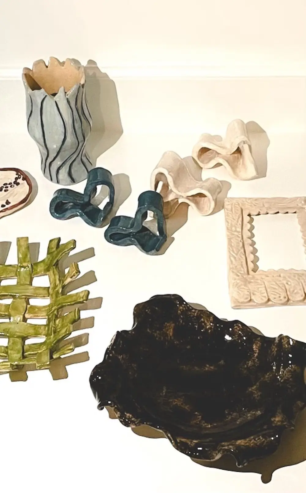 O'More students' pottery: A collection of ceramic objects displayed on a white surface, including a wavy-edged vase with black line details, abstract sculptural pieces in cream and deep blue, a textured picture frame, and a variety of uniquely shaped decorative pieces in green, black, and neutral tones.