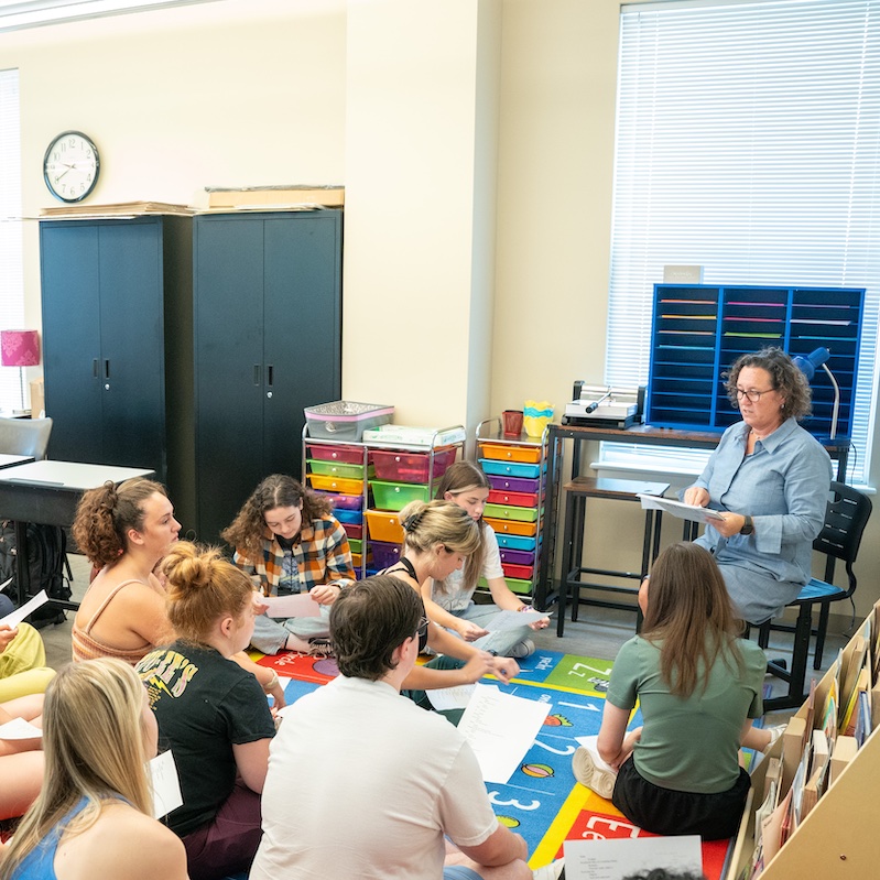 The image shows a classroom setting with a group of students sitting at desks and focused on colorful worksheets or papers. An instructor, who appears to be a woman with glasses and curly hair, stands at the front of the classroom near a whiteboard. The classroom has typical educational furniture including student desks arranged in rows, storage cabinets along the walls, colorful organizational drawers, and a wall clock. Natural light comes in through windows with blinds. The students appear engaged in their classwork while the instructor seems to be providing guidance or instruction.