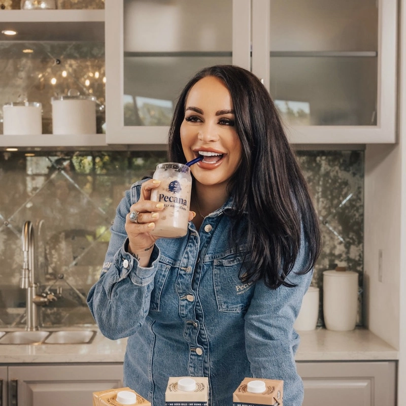 A woman smiling in a kitchen, holding a glass with a straw that has 'Pecana' on it, wearing a denim jacket, with containers of Pecana products visible on the counter.
