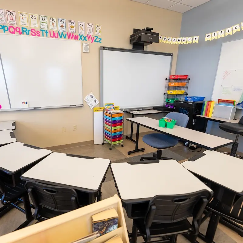 A small classroom with desks arranged in a semicircle formation. The room features whiteboards mounted on the walls, an alphabet display across the top of one wall, and a projector mounted to the ceiling. Colorful organizational elements include plastic storage bins in various colors, a rainbow-colored rolling cart with drawers, and some educational materials. The classroom has chairs for students, a teacher's desk area, and appears designed for elementary education with its bright, organized layout.