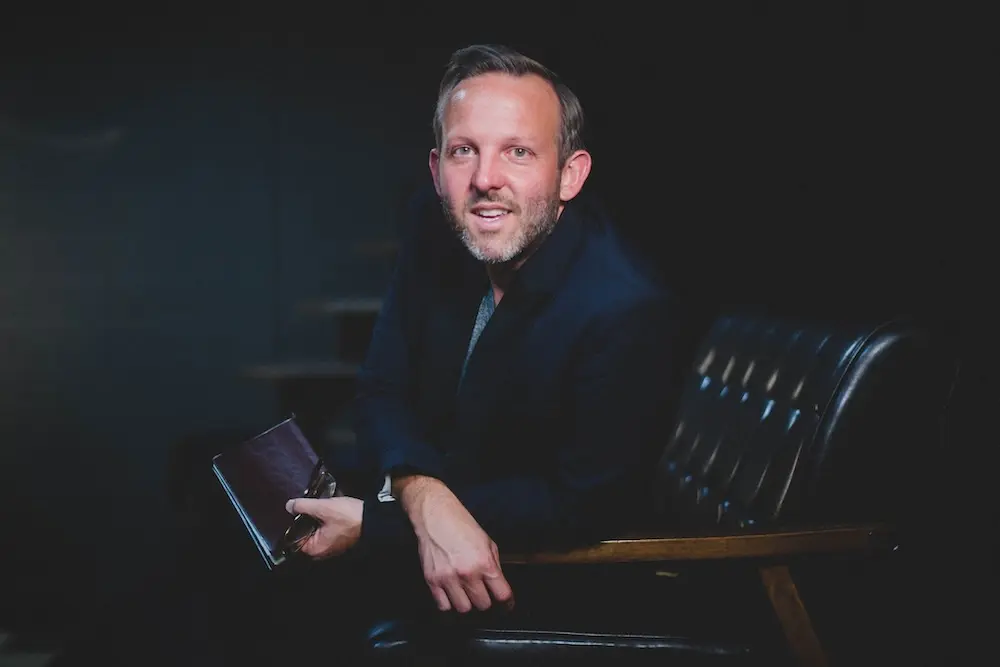 Greer sitting on a sofa leaning toward the camera