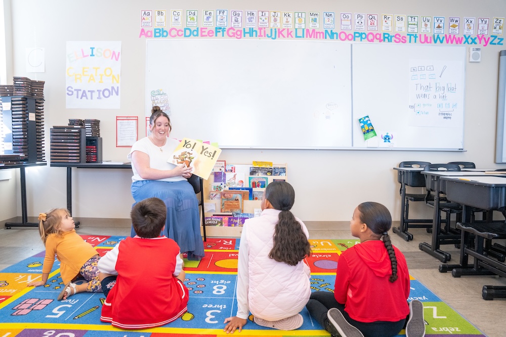 A smiling teacher sits in a chair holding up a picture book titled "Yo! Yes!" to four young children seated on a colorful alphabet-and-number rug. A reading shelf, an alphabet banner, and a whiteboard with a handwritten sentence are visible in the classroom behind them.