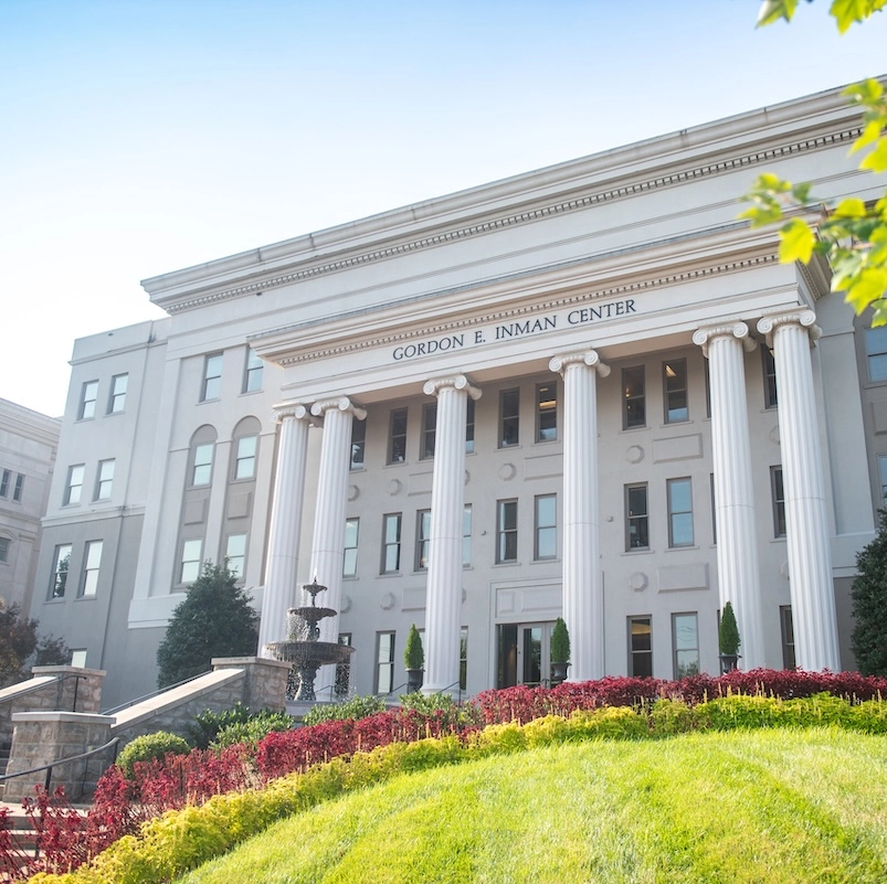 Exterior view of the Gordon E. Inman Center, a white neoclassical building with prominent columns, surrounded by red flowering plants and green landscaping on a hillside.