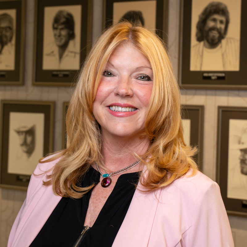 Smiling woman with long blonde hair wearing a pink blazer, against a background of framed portraits.
