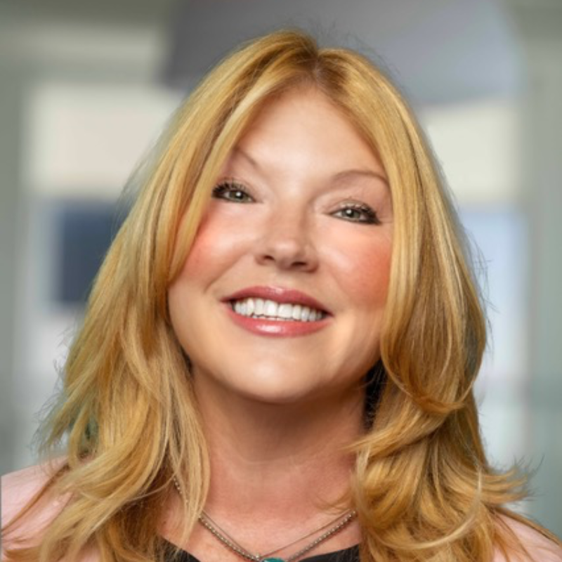 Jodi Marr smiling with long blonde hair wearing a pink blazer, against a background of framed portraits.