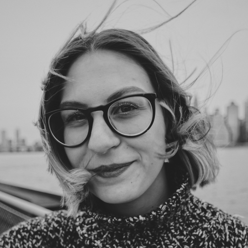 Smiling woman with glasses and wind-blown hair in a black and white portrait, city skyline in the background.