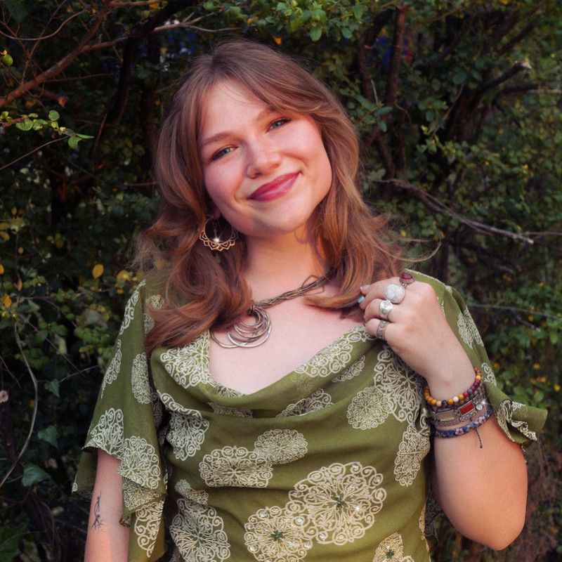 Smiling young woman with long hair, wearing a green patterned top, layered necklace, rings, and bracelets outdoors.