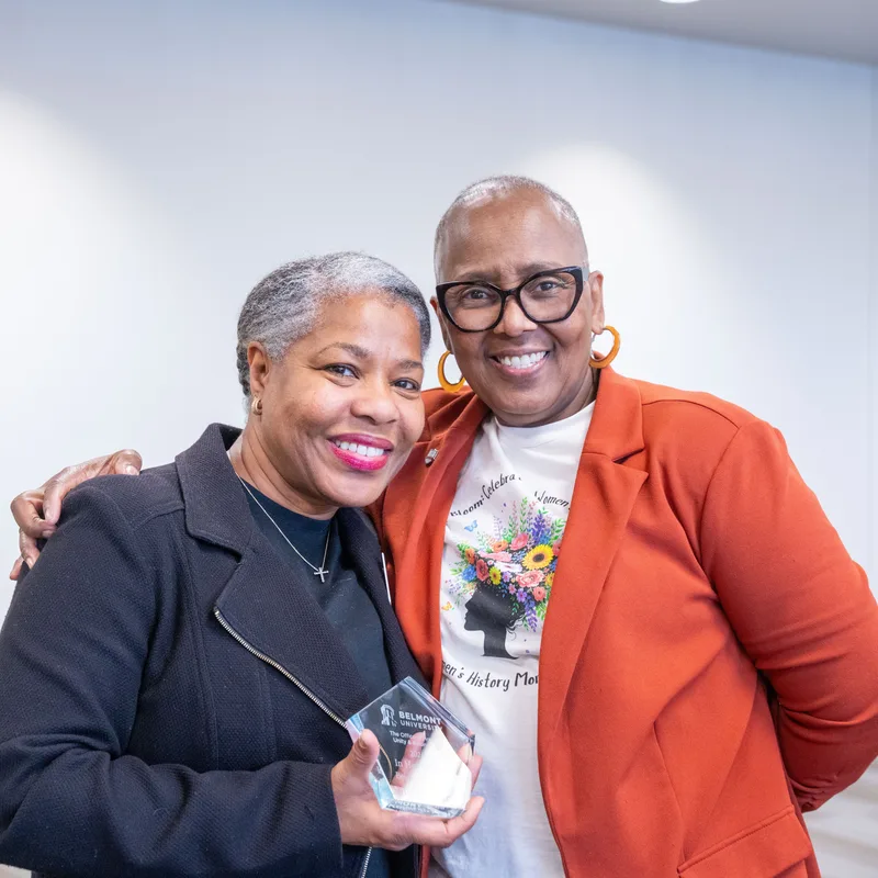 Two women smiling together at an indoor event. The woman on the left wears a dark navy blazer and holds a crystal award. The woman on the right wears a bright orange cardigan over a graphic t-shirt and glasses. Both have short gray hair and are embracing warmly in front of a light blue interior background.