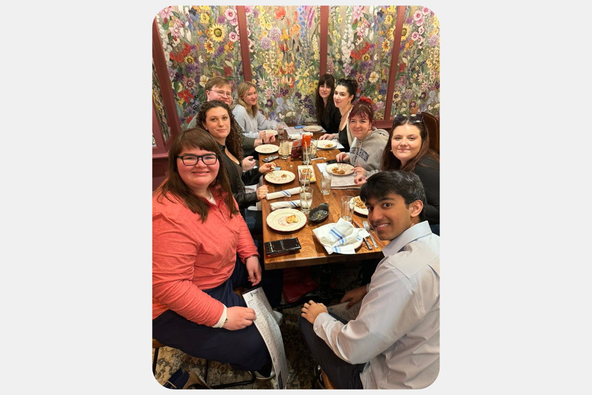 A group of friends smiling while dining together at a restaurant with a vibrant floral wallpaper background.