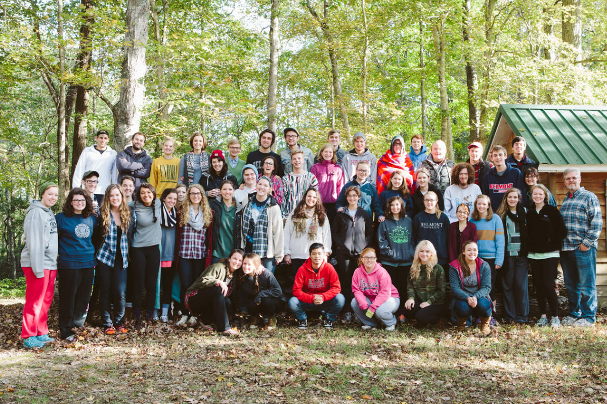 Diverse group of young adults and mentors smiling in a forest setting with a rustic cabin, ideal for youth camp or retreat.
