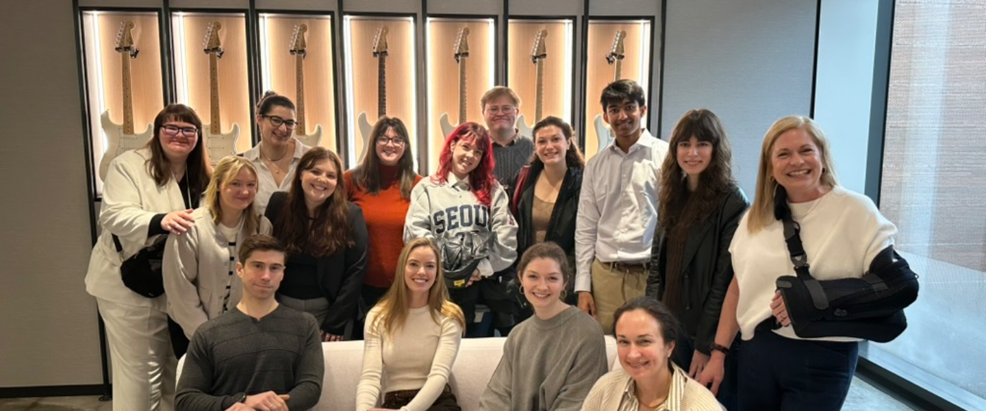 Smiling group of students and adults in a room with a display of electric guitars on the wall.