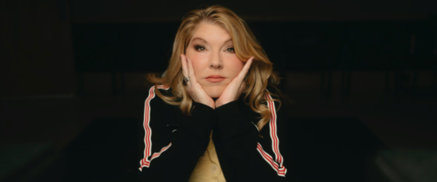Blonde woman in black track jacket with red and white stripes, hands cupping face, pensive expression.