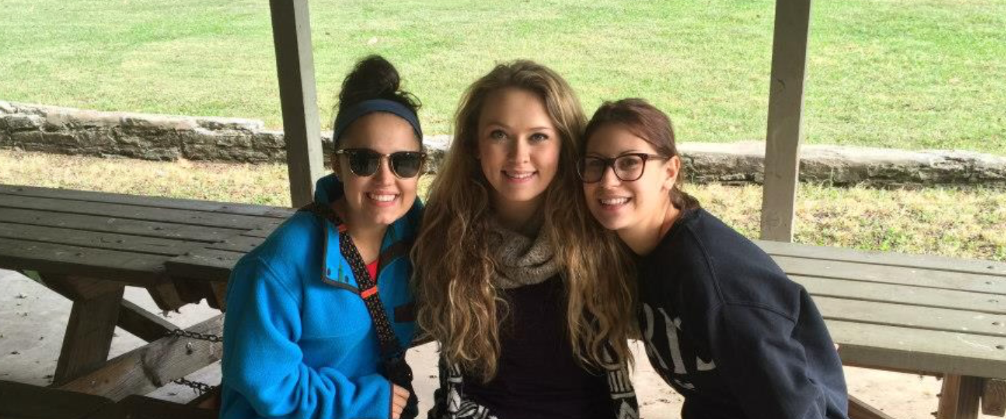 Three happy young women sitting on a wooden picnic table under a park shelter, smiling at the camera.