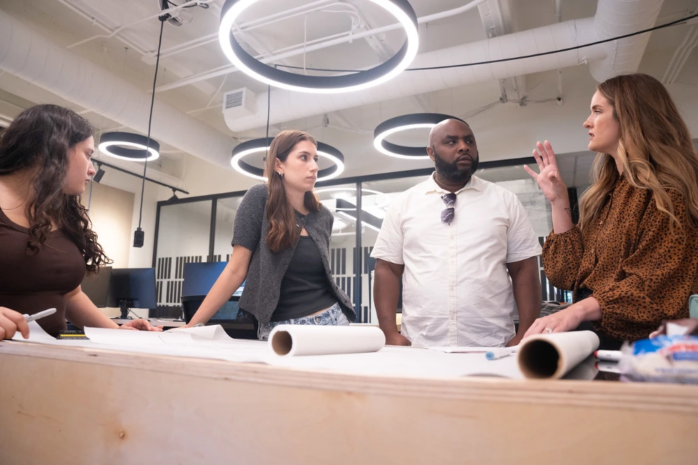Students and faculty discussing architectural plans at a standing desk in a modern office.
