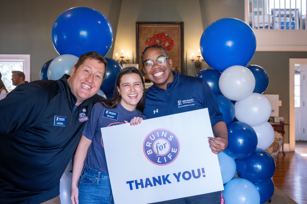 Three smiling Belmont University members hold a 'Bruins for Life, Thank You!' sign with blue and white balloons.