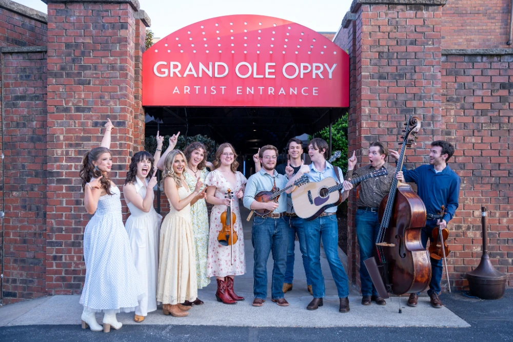 Bluegrass and country musicians pose with instruments in front of the Grand Ole Opry Artist Entrance sign.