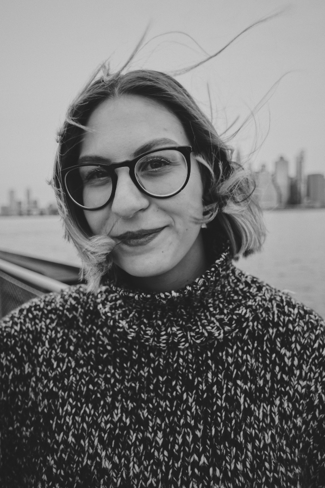 Smiling woman with glasses and wind-blown hair in a black and white portrait, city skyline in the background.