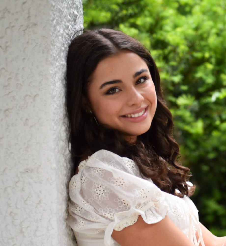 Portrait of a happy young woman with dark wavy hair wearing a white top, smiling outdoors against a textured wall.
