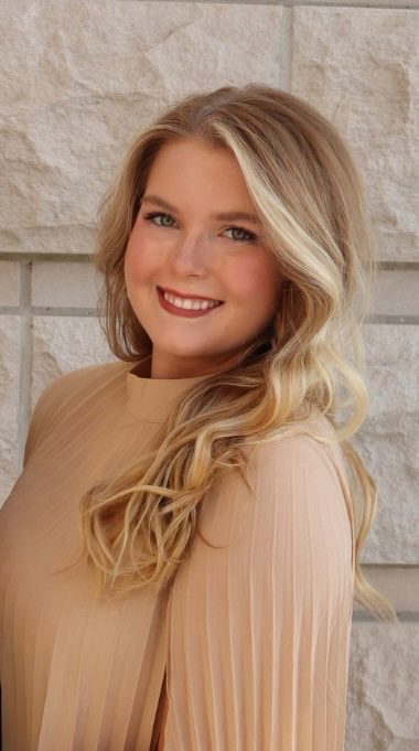 Smiling young woman with blonde hair wearing a beige pleated top against a light stone background.