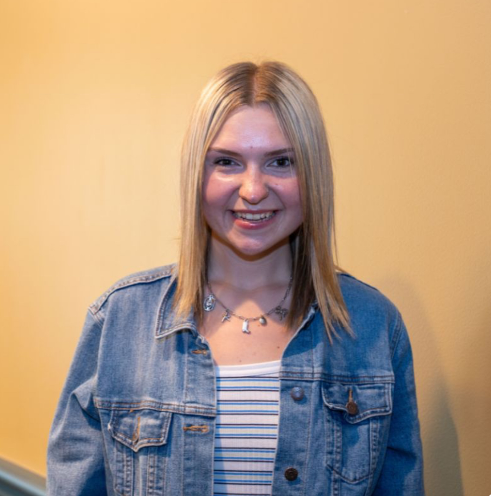 Smiling young woman with blonde hair in a denim jacket and striped top, wearing a charm necklace.