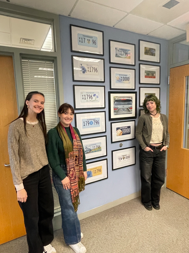 Three women smiling by a wall of framed Tennessee 1796 commemorative license plates and historical art.