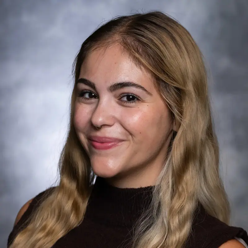 Studio headshot of a smiling young woman with long wavy blonde hair against a gray background.