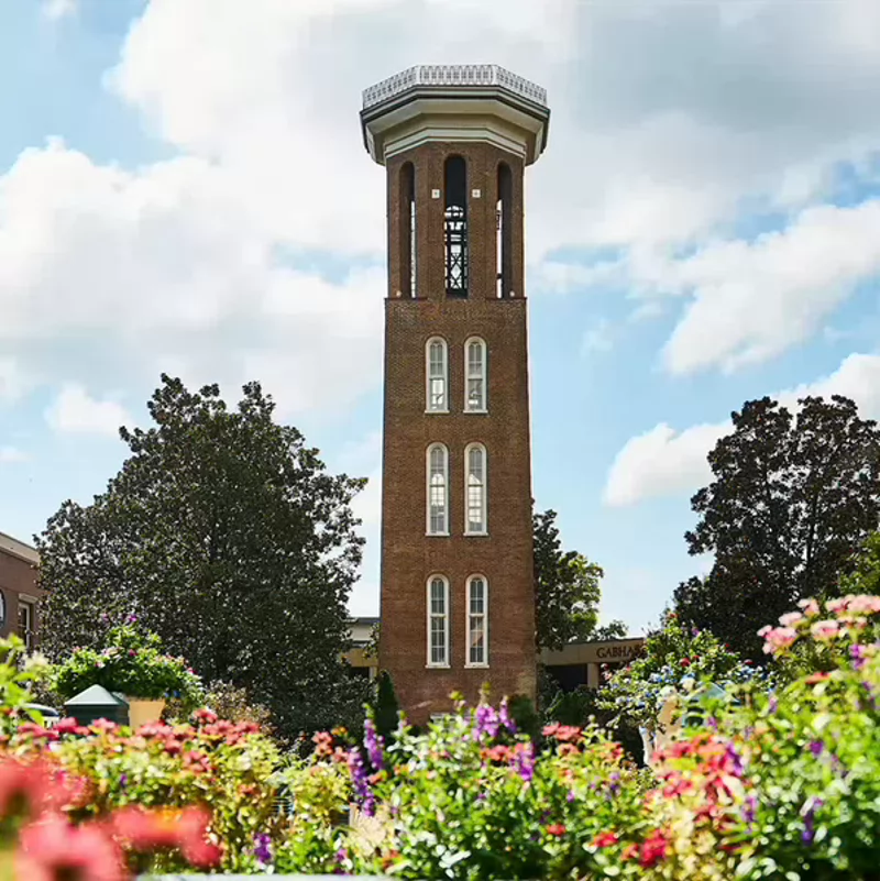 Belmont Bell Tower rises above colorful flower gardens on a bright spring day with blue skies and scattered clouds.