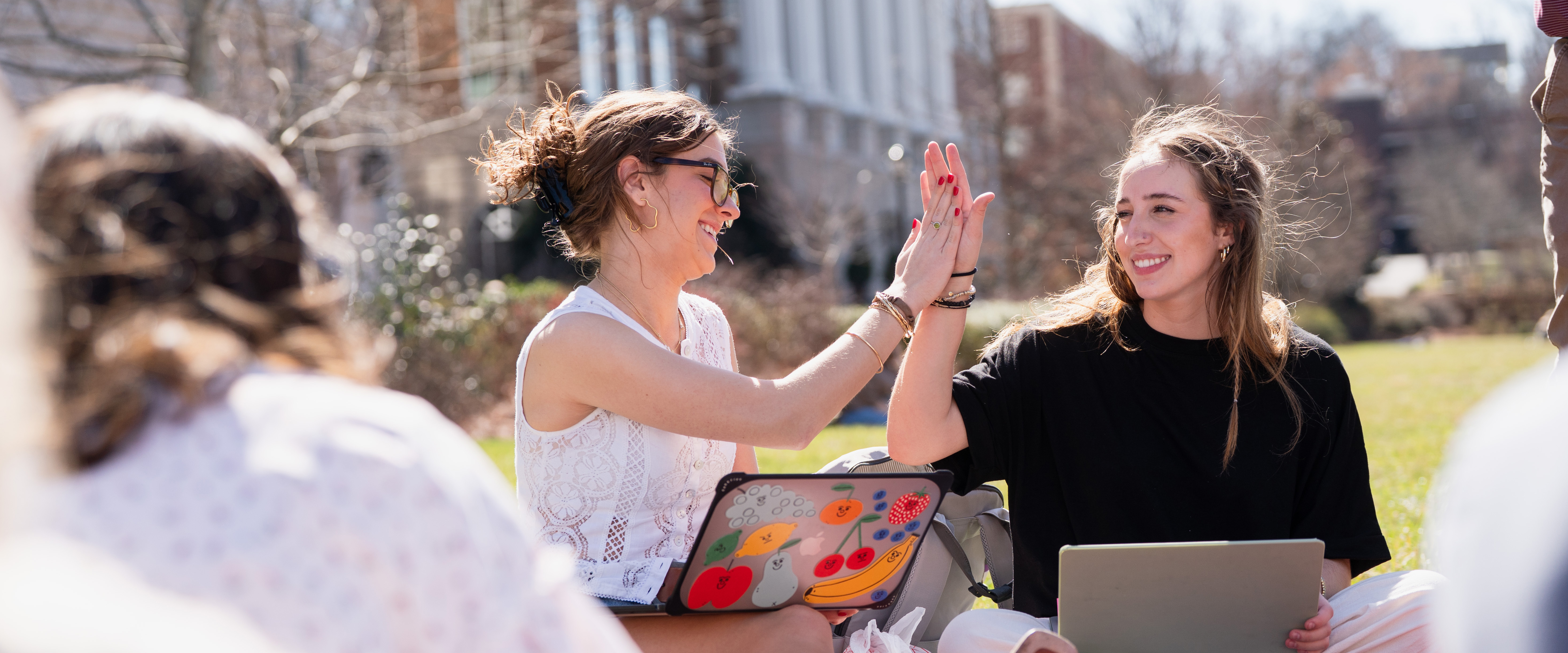Students sitting on the lawn on a sunny day high fiving with laptops in their laps