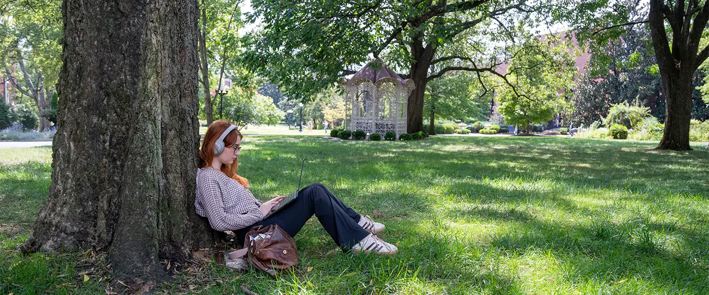 A student with headphones sits against a large tree, working on a laptop in a serene area of Belmont University’s campus, with the gazebo visible in the background.