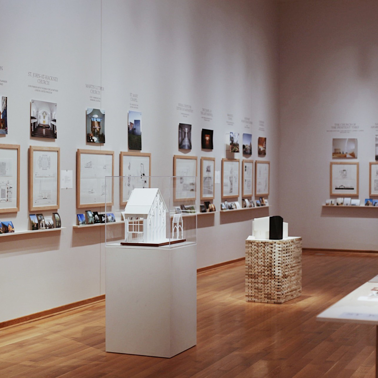 a white walled gallery has architectural images hanging with models in the center of the room