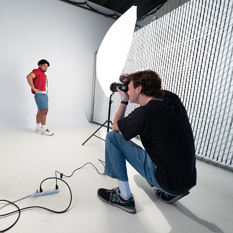 Image of one teen pointing a camera at another teen in front of a backdrop with an umbrella light. 