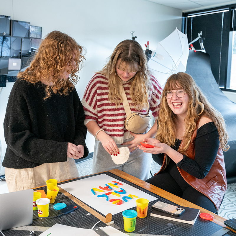 Three women sitting around a table creating handmade letters out of playdough while laughing. 