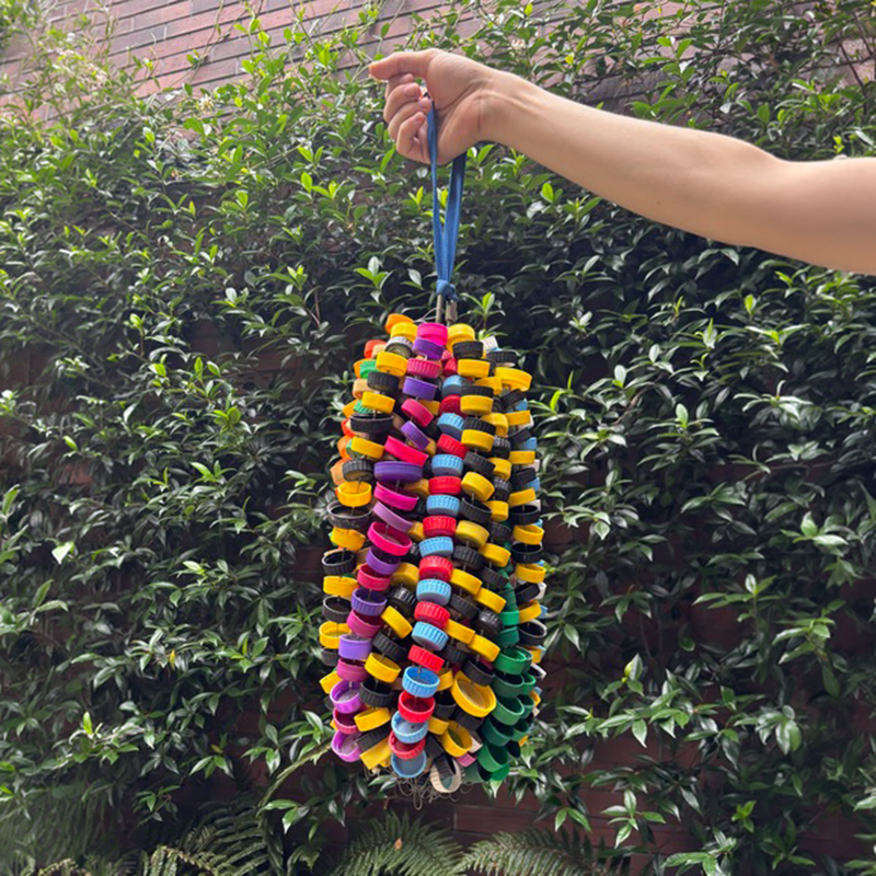 an outstretched arm holding a hanging sculpture made of different colored plastic bottle caps in front of greenery